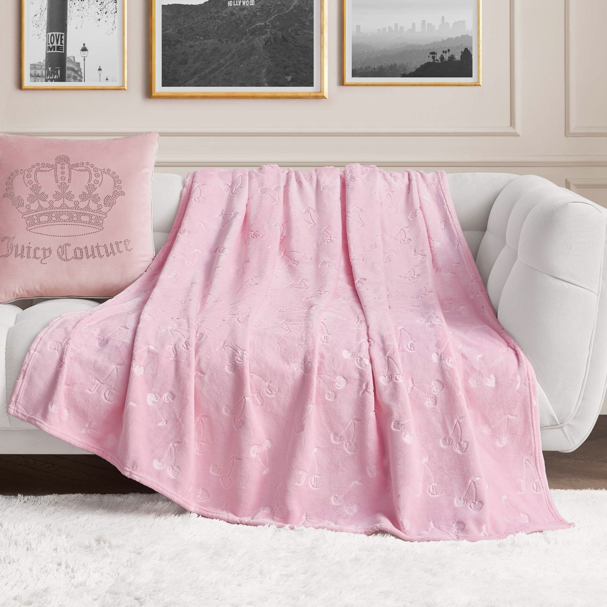 An Embossed Plush Oversized Throw Blanket in light pink from Juicy Couture is draped over a white couch, paired with a matching pink pillow and framed wall art for a cozy, stylish living room.