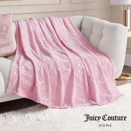 An Embossed Plush Oversized Throw Blanket in light pink is draped over a white sofa, with a matching pillow and decor behind it. "Juicy Couture Home" is displayed in the bottom right corner.