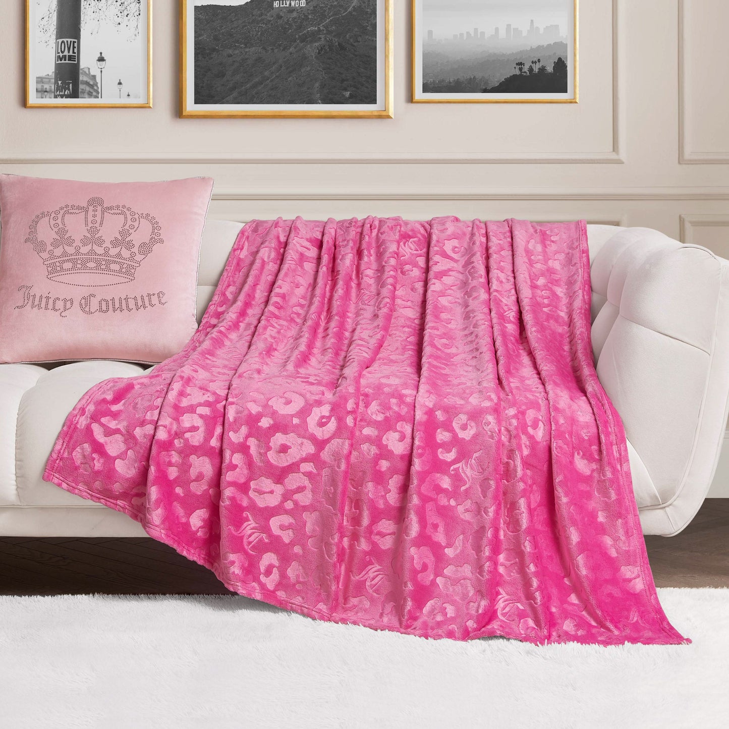 An Embossed Plush Oversized Throw Blanket in pink leopard print is draped over a white sofa, paired with a light pink "Juicy Couture" pillow. Framed photos and crisp white paneled walls complete the chic scene.