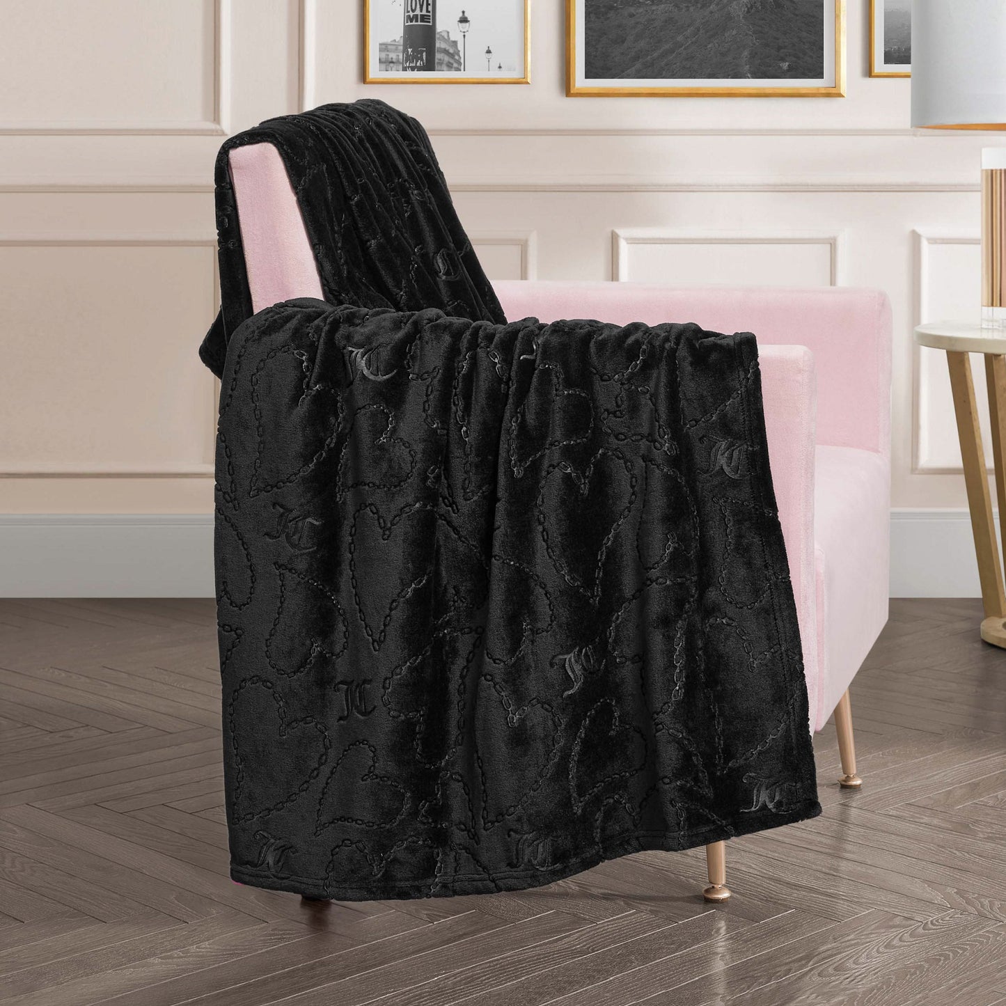 The Embossed Plush Oversized Throw Blanket in black, featuring a subtle heart pattern, is draped over a light pink armchair in a modern living room with framed art and a round side table.