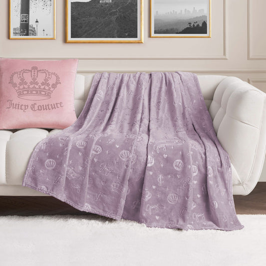 An Embossed Plush Oversized Throw Blanket in light purple with a crown and logo pattern is draped over a white sofa, paired with a pink Juicy Couture pillow in a stylish living room featuring framed wall art.