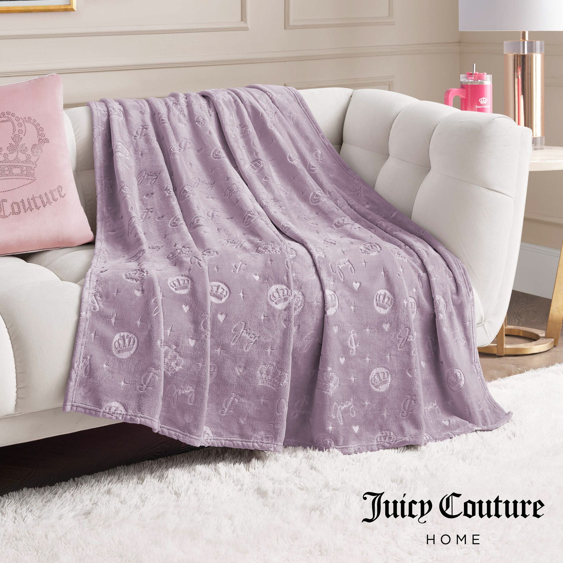 An Embossed Plush Oversized Throw Blanket in lavender is draped over a white sofa, paired with a matching pink Juicy Couture pillow. The elegant room decor and Juicy Couture Home text appear in the bottom right corner.