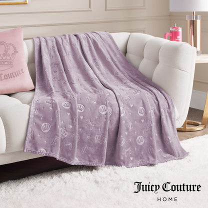 An Embossed Plush Oversized Throw Blanket in lavender is draped over a white sofa, paired with a matching pink Juicy Couture pillow. The elegant room decor and Juicy Couture Home text appear in the bottom right corner.