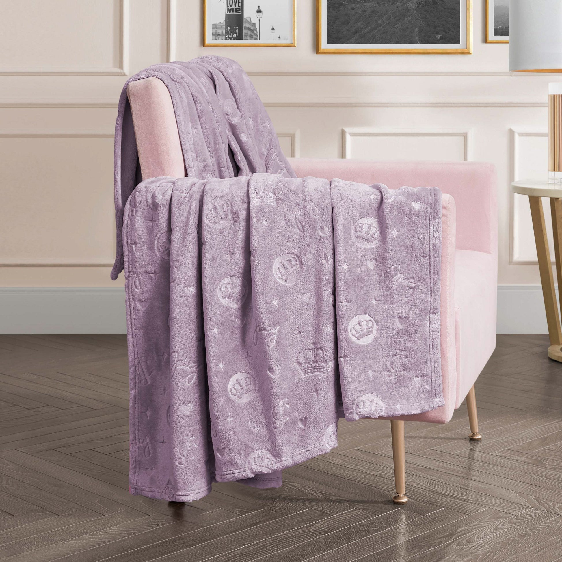 An Embossed Plush Oversized Throw Blanket with crown and heart patterns in light purple is draped over a light pink armchair in a modern living room featuring wood flooring and framed wall art.