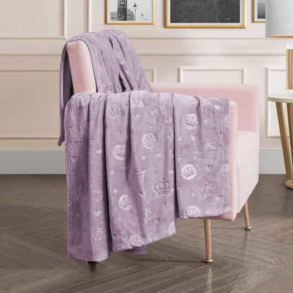 An Embossed Plush Oversized Throw Blanket with crown and heart patterns in light purple is draped over a light pink armchair in a modern living room featuring wood flooring and framed wall art.