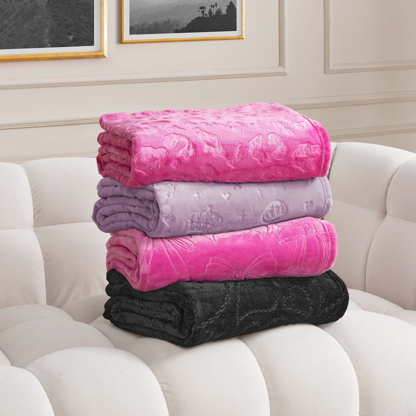 Four Embossed Plush Oversized Throw Blankets in pink, lavender, magenta, and black are stacked on a white tufted sofa in a room with framed wall art and light paneled walls.