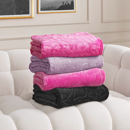 Four Embossed Plush Oversized Throw Blankets in pink, lavender, magenta, and black are stacked on a white tufted sofa in a room with framed wall art and light paneled walls.