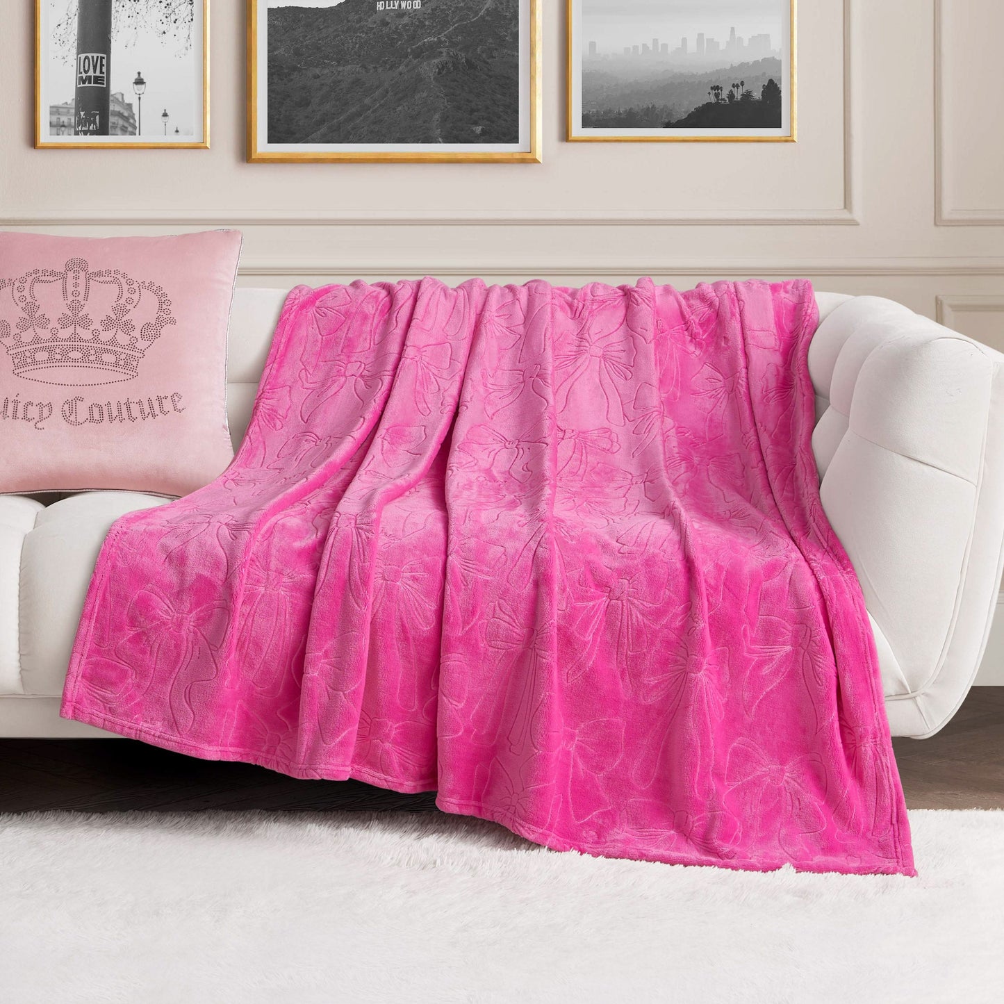 An Embossed Plush Oversized Throw Blanket in bright pink is draped over a white sofa. A light pink pillow with a crown design rests nearby, while framed artwork decorates the wall behind.