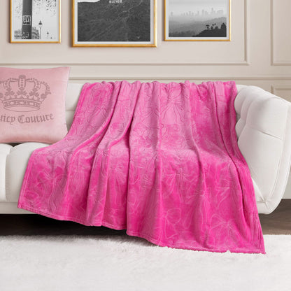 An Embossed Plush Oversized Throw Blanket in bright pink is draped over a white sofa. A light pink pillow with a crown design rests nearby, while framed artwork decorates the wall behind.