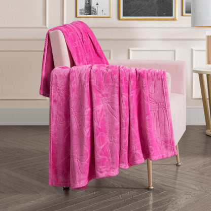 An Embossed Plush Oversized Throw Blanket in bright pink, featuring a bow pattern, is draped over a light pink upholstered chair in a modern living room with wood flooring and framed art on the wall.