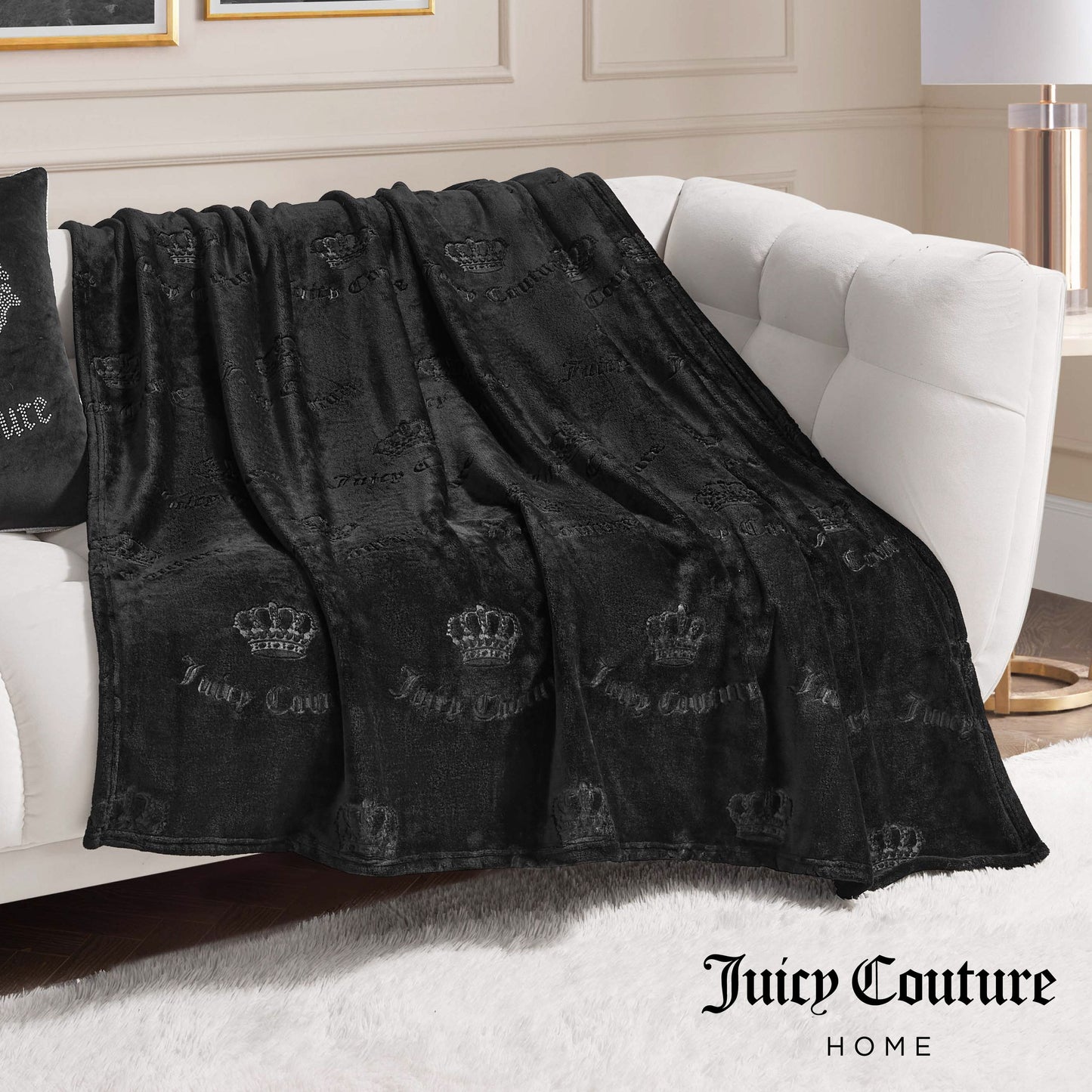 An Embossed Plush Oversized Throw Blanket by Juicy Couture, featuring a crown and logo pattern, is draped over a white sofa. The soft black blanket displays the Juicy Couture Home logo in the bottom right corner of the image.