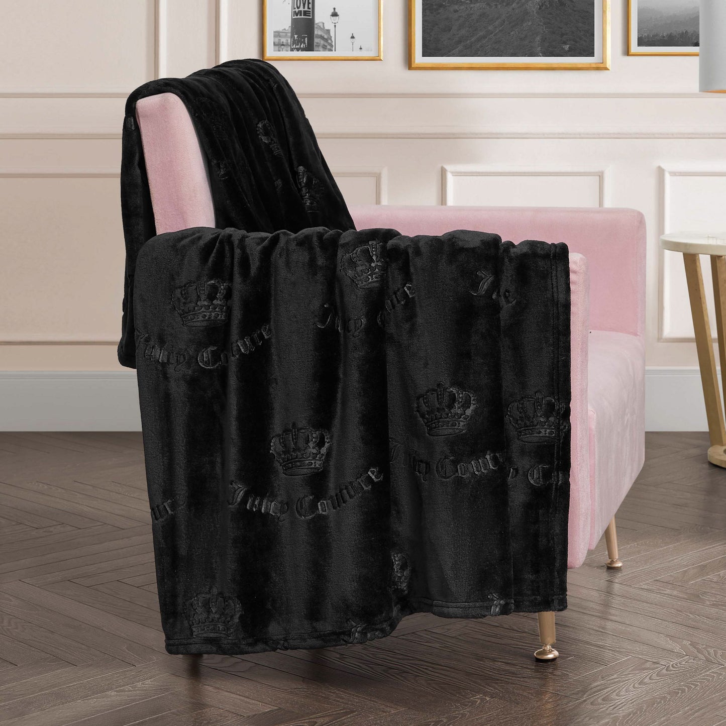 An Embossed Plush Oversized Throw Blanket from Juicy Couture with a crown pattern is draped over a light pink armchair in a chic modern room with white paneled walls, framed art, and a wooden herringbone floor.