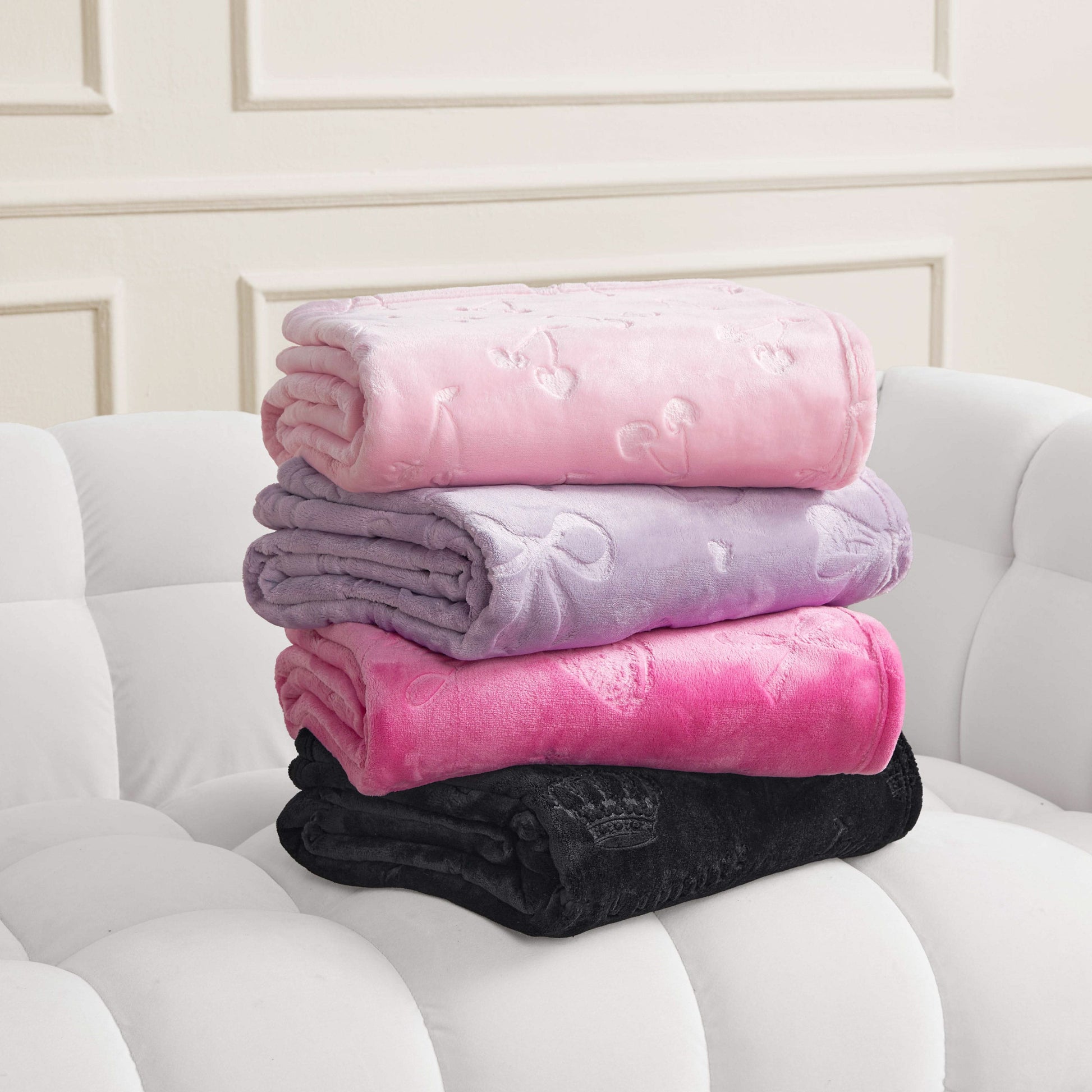 Four Embossed Plush Oversized Throw Blankets in light pink, lavender, hot pink, and black are stacked on a white tufted sofa; each soft blanket features a subtle bow pattern, adding charm against a cream wall.
