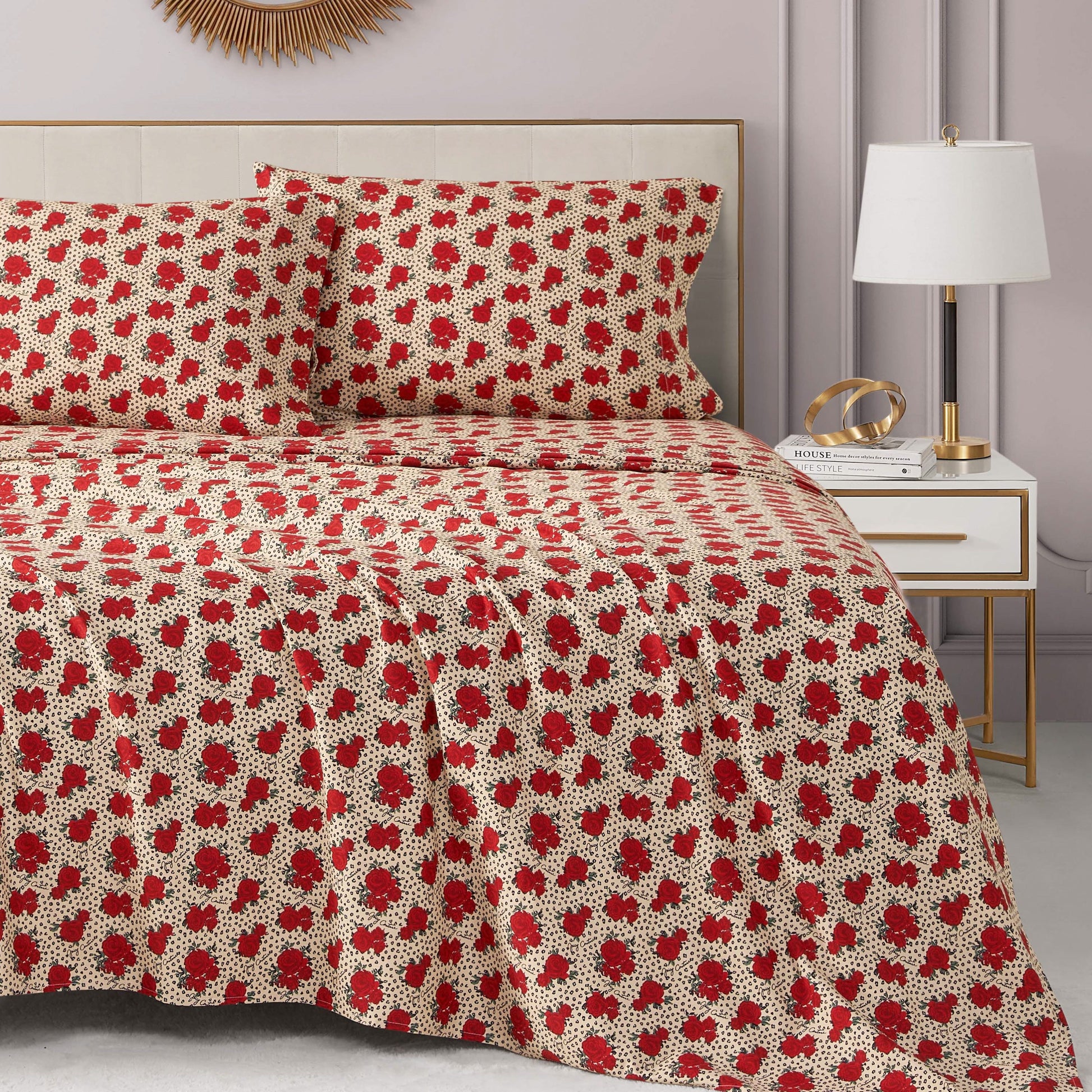 A neatly made bed with a red and white floral Printed Microfiber Sheet Set and matching pillows. The room has a modern nightstand with a white lamp, books, gold decor, and a sunburst mirror on a light-colored wall.