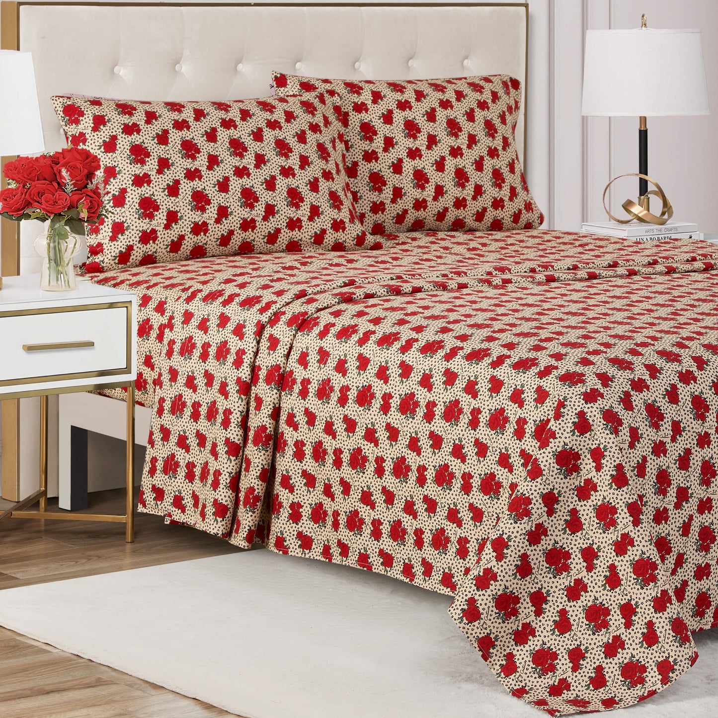A neatly made bed with a cream and red floral-patterned Printed Microfiber Sheet Set and matching pillowcases, flanked by white nightstands with lamps and a vase of red roses. The room features modern decor and a tufted headboard.