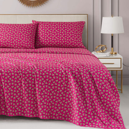 A neatly made bed with a bright pink Printed Microfiber Sheet Set and matching pillowcases featuring a small leafy pattern. The room includes a white nightstand, lamp, gold décor, and neutral walls for an added touch of luxury.