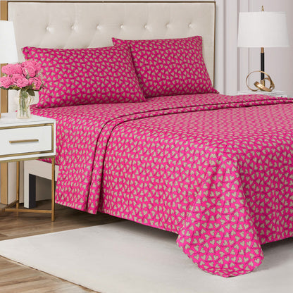 A neatly made bed with a pink and gold patterned bedspread and matching pillowcases sits against a tufted beige headboard. A Printed Microfiber Sheet Set adds elegance, paired with a nightstand holding pink flowers and a lamp.