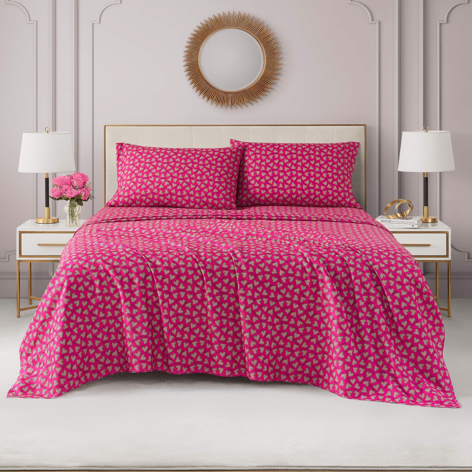 A neatly made bed with a bright pink Printed Microfiber Sheet Set and matching pillows featuring a small geometric pattern, flanked by white nightstands with lamps, flowers, and a round sunburst mirror on a pale pink wall.