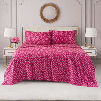 A neatly made bed with a bright pink Printed Microfiber Sheet Set and matching pillows featuring a small geometric pattern, flanked by white nightstands with lamps, flowers, and a round sunburst mirror on a pale pink wall.