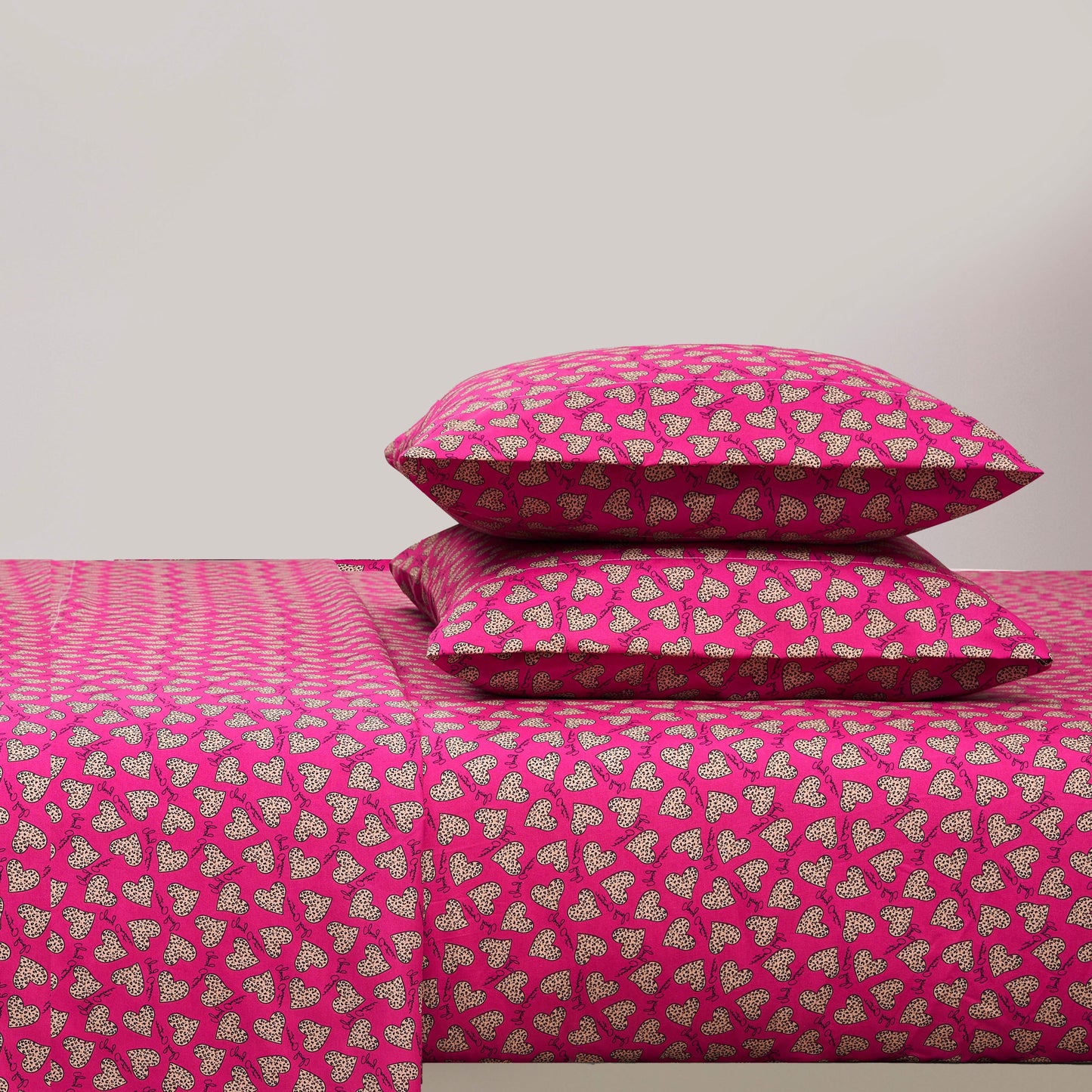 A bed with a bright pink Printed Microfiber Sheet Set and two matching pillows, all featuring a repeating pattern of small white and black hearts. The background wall is plain and light-colored.