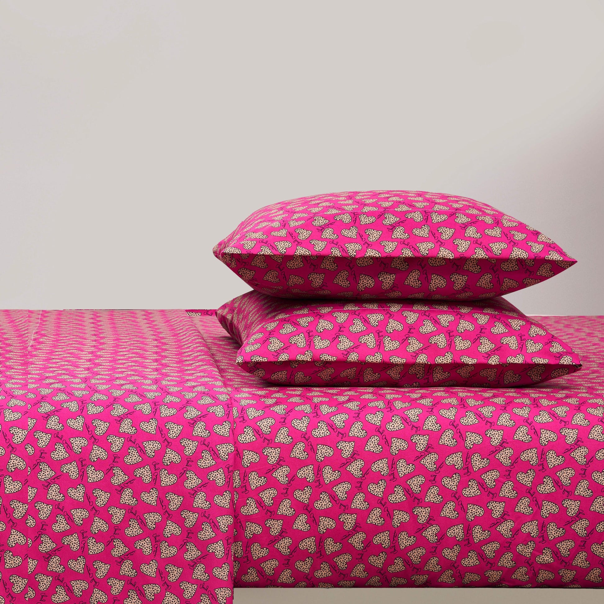 A bed with a bright pink Printed Microfiber Sheet Set and two matching pillows, all featuring a repeating pattern of small white and black hearts. The background wall is plain and light-colored.