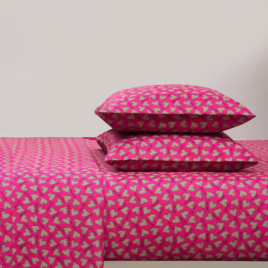 A bed with a bright pink Printed Microfiber Sheet Set and two matching pillows, all featuring a repeating pattern of small white and black hearts. The background wall is plain and light-colored.