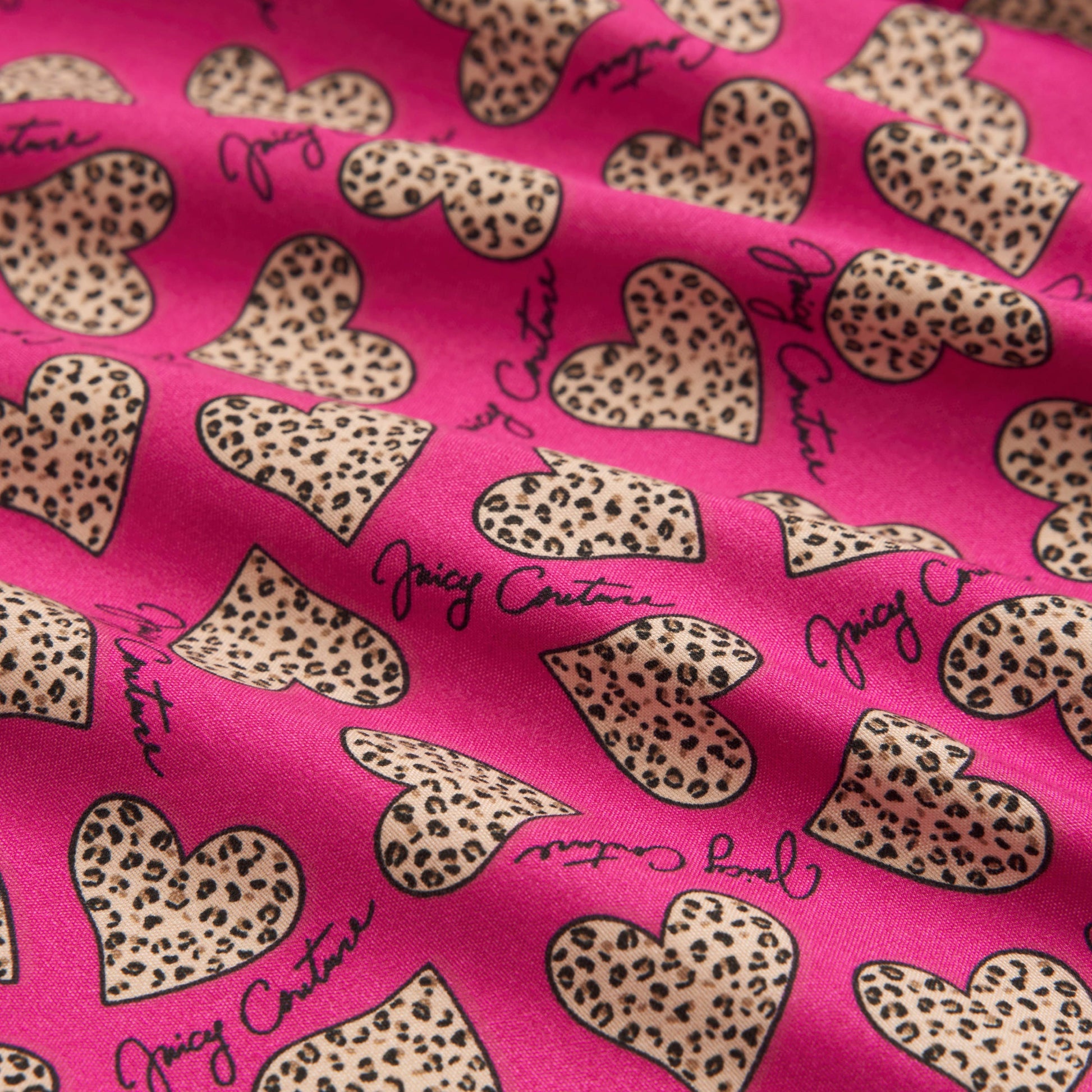 A close-up of pink fabric from the Printed Microfiber Sheet Set shows leopard-print hearts and "Juicy Couture" in black script. The slightly wrinkled microfiber gives this luxury bedding a relaxed, stylish appearance.