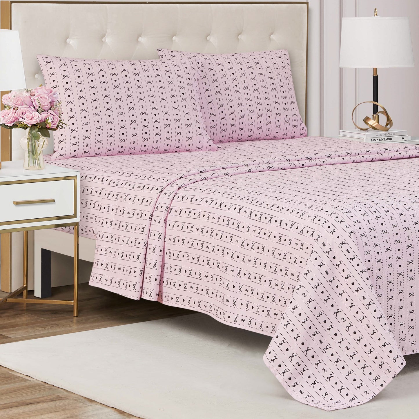 A neatly made bed with a Printed Microfiber Sheet Set in pink patterns and two matching pillows stands in a stylish bedroom. A nightstand with pink flowers, a lamp, and books adds to the modern, elegant luxury bedding decor.
