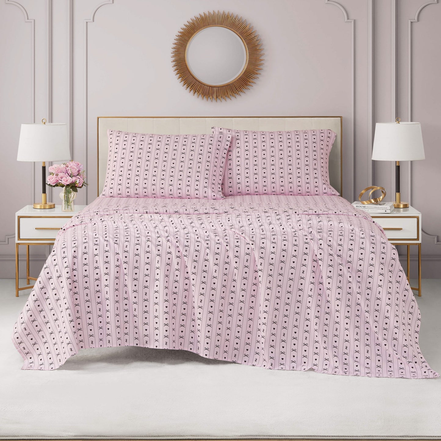 A neatly made bed with a pink-patterned Printed Microfiber Sheet Set and two pillows, flanked by matching white nightstands with lamps and flowers. A round gold sunburst mirror hangs above the bed against a light gray wall.