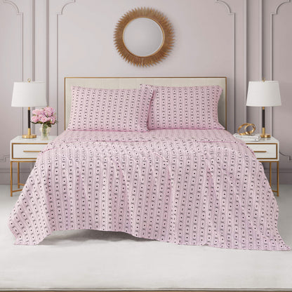 A neatly made bed with a pink-patterned Printed Microfiber Sheet Set and two pillows, flanked by matching white nightstands with lamps and flowers. A round gold sunburst mirror hangs above the bed against a light gray wall.