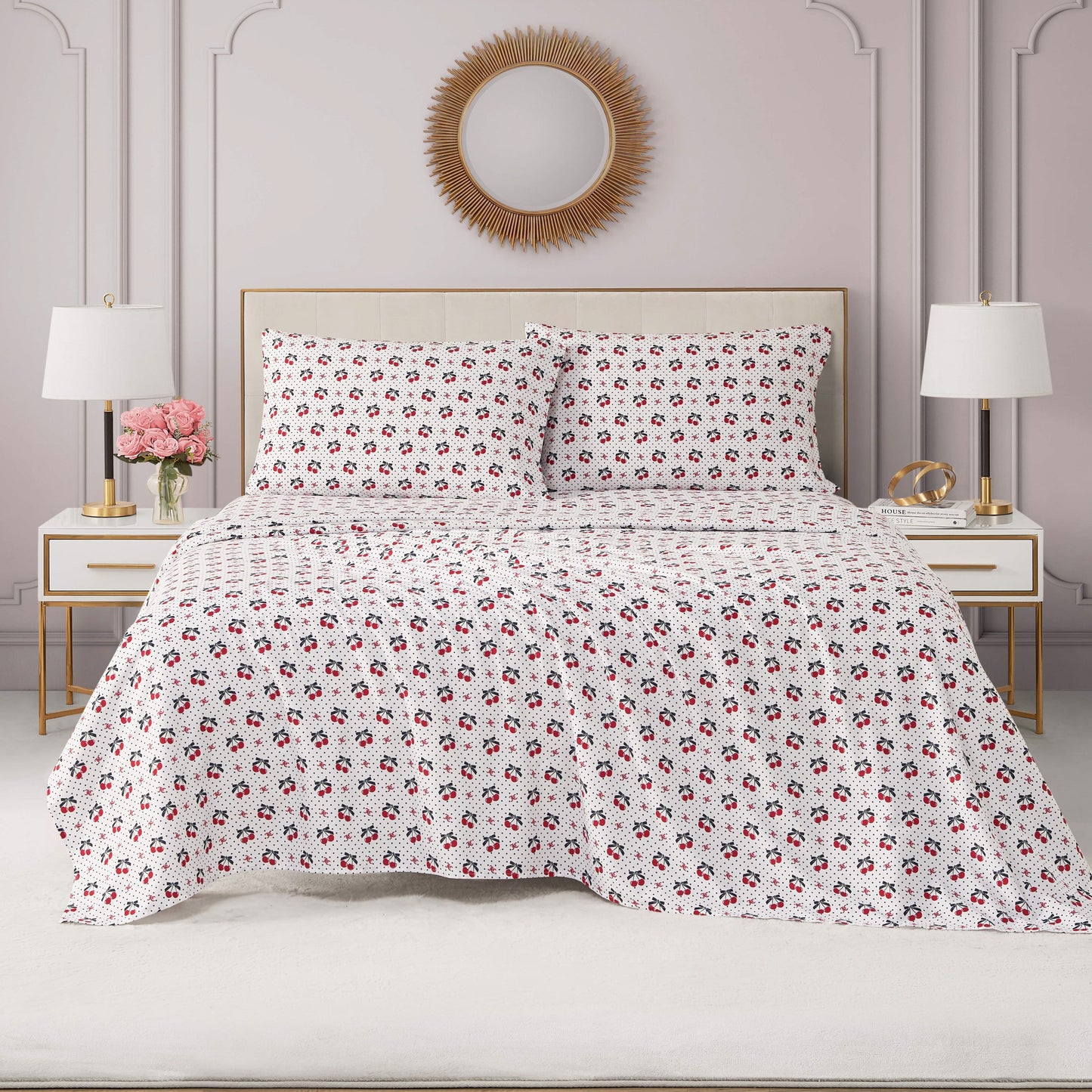 A neatly made bed with a white and red floral quilt, a Printed Microfiber Sheet Set, matching pillows, white nightstands with gold accents, a vase of pink flowers, and a sunburst mirror on a light pink wall.