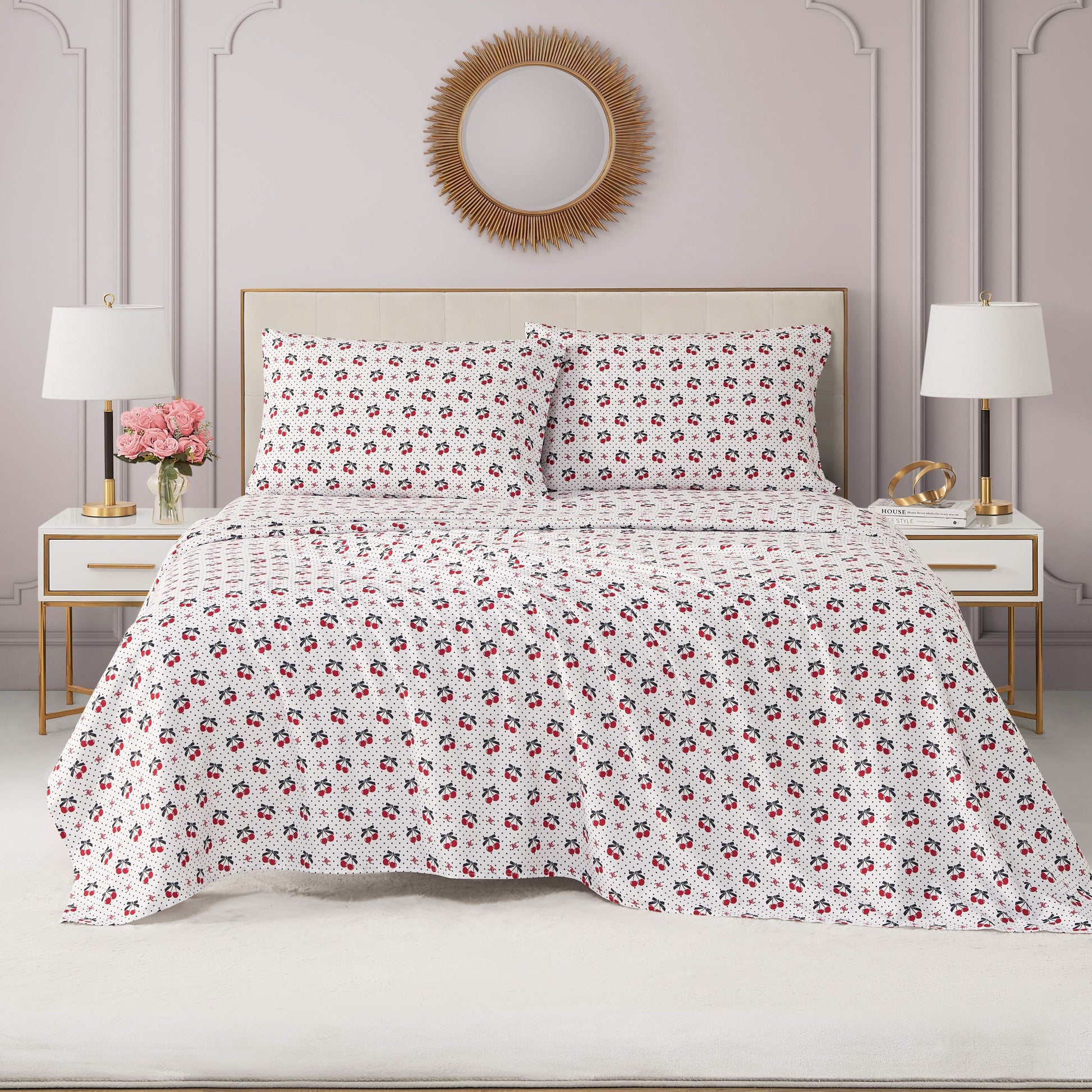 A neatly made bed with a white and red floral quilt, a Printed Microfiber Sheet Set, matching pillows, white nightstands with gold accents, a vase of pink flowers, and a sunburst mirror on a light pink wall.