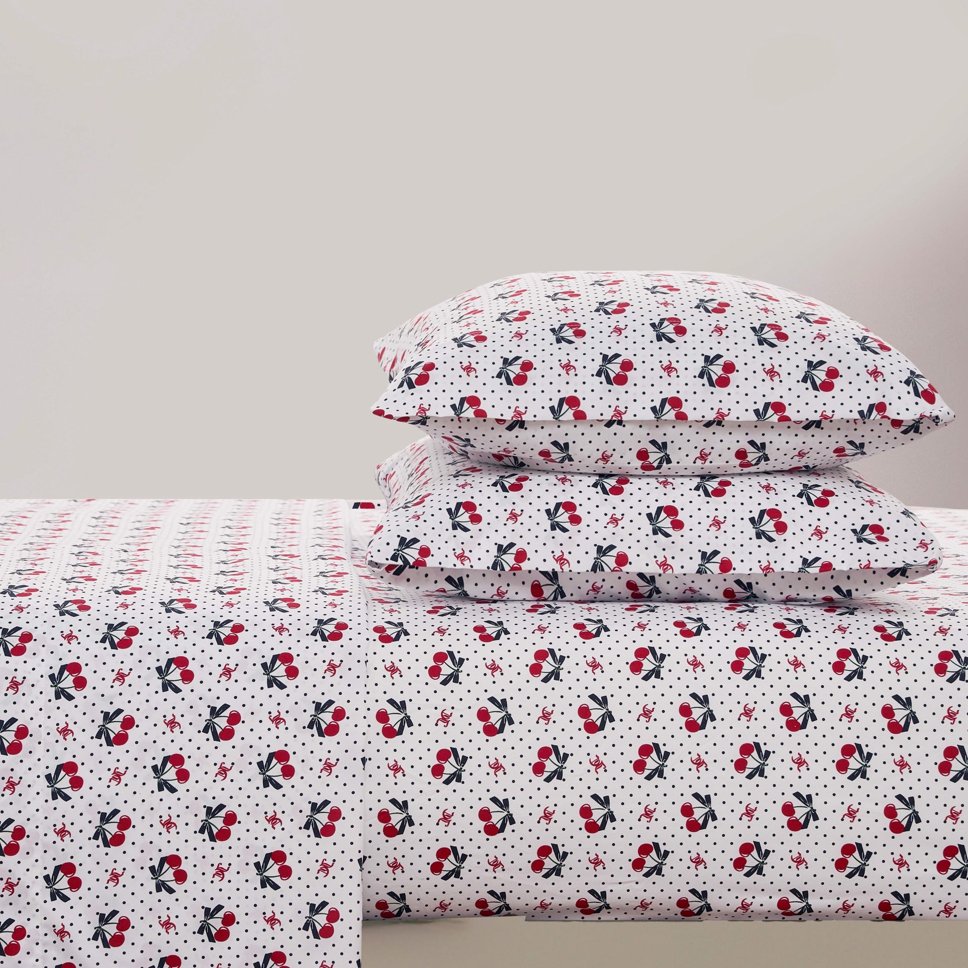 A neatly made bed with a Printed Microfiber Sheet Set, including pillowcases and a blanket decorated with small red flowers, black leaves, and scattered black polka dots on a plain light background.