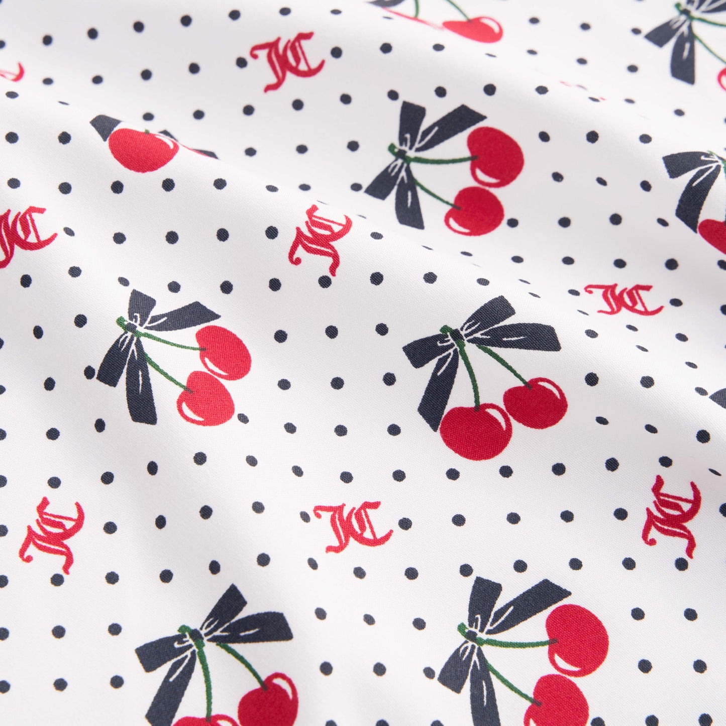 The Printed Microfiber Sheet Set features white fabric with small black polka dots, red cherries tied with black bows, and scattered red KC initials for a playful Juicy Couture touch. Slightly wrinkled for relaxed luxury bedding style.