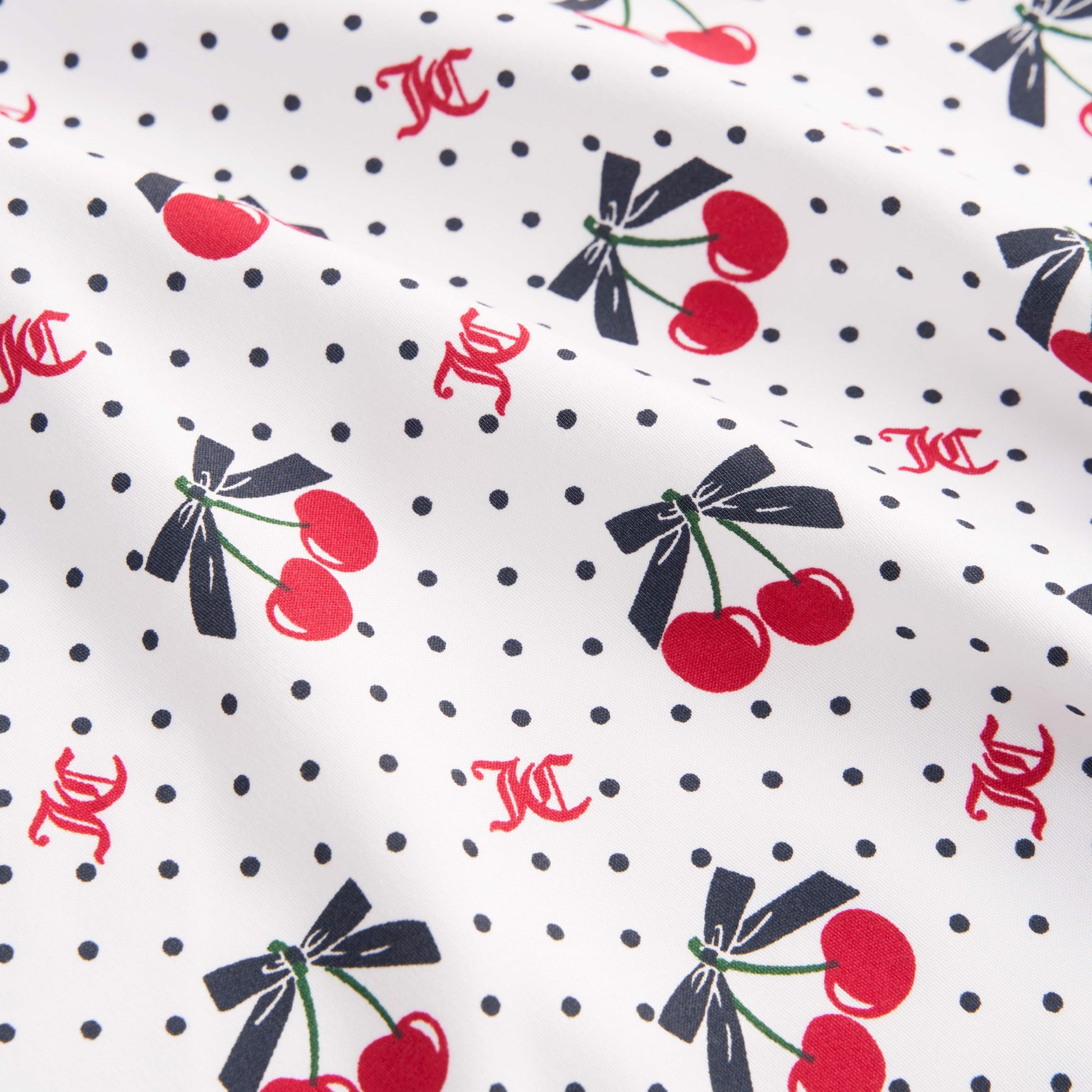 The Printed Microfiber Sheet Set features white fabric with small black polka dots, red cherries tied with black bows, and scattered red KC initials for a playful Juicy Couture touch. Slightly wrinkled for relaxed luxury bedding style.