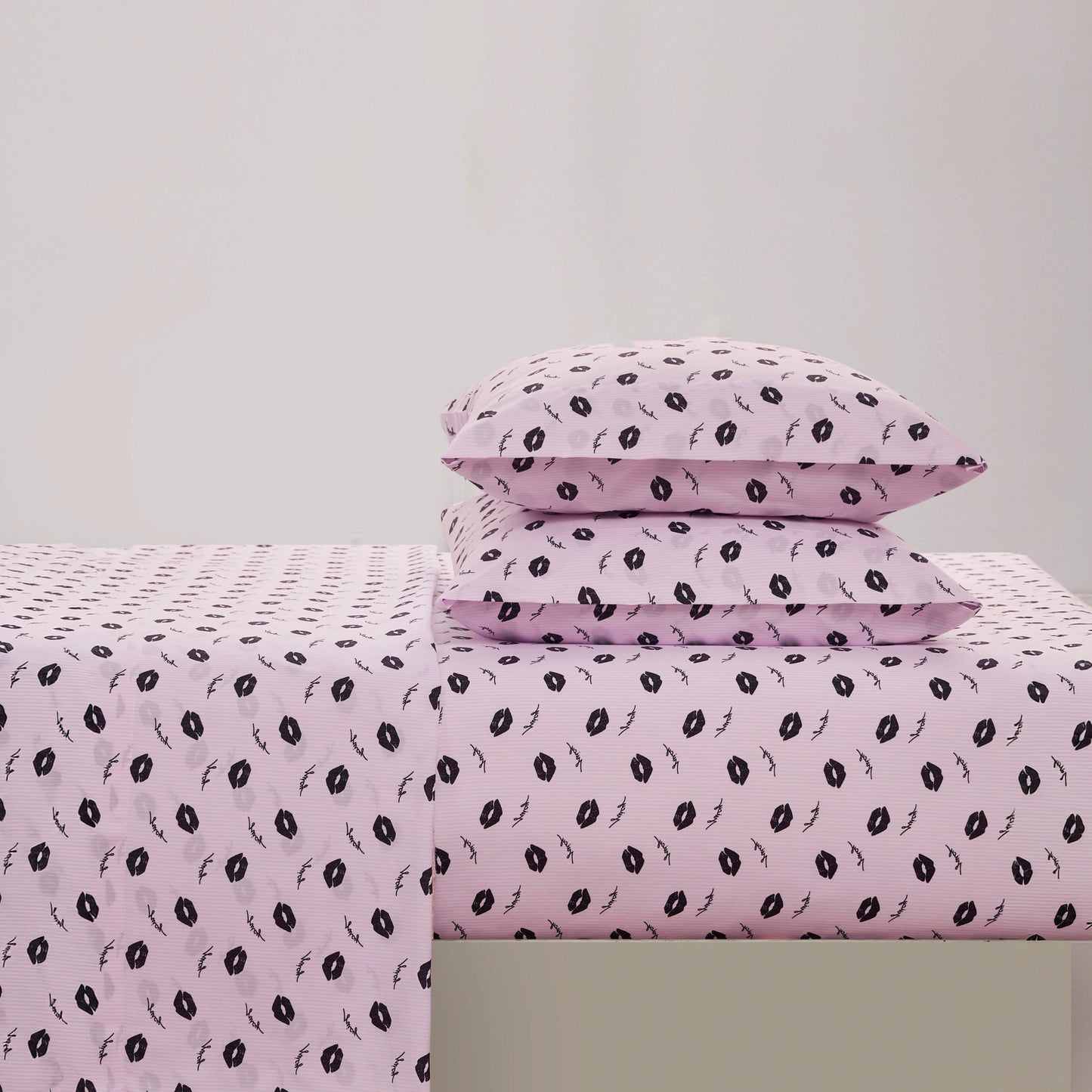 A neatly made bed with a light pink Printed Microfiber Sheet Set featuring black lips print pillowcases, set against a plain white background.