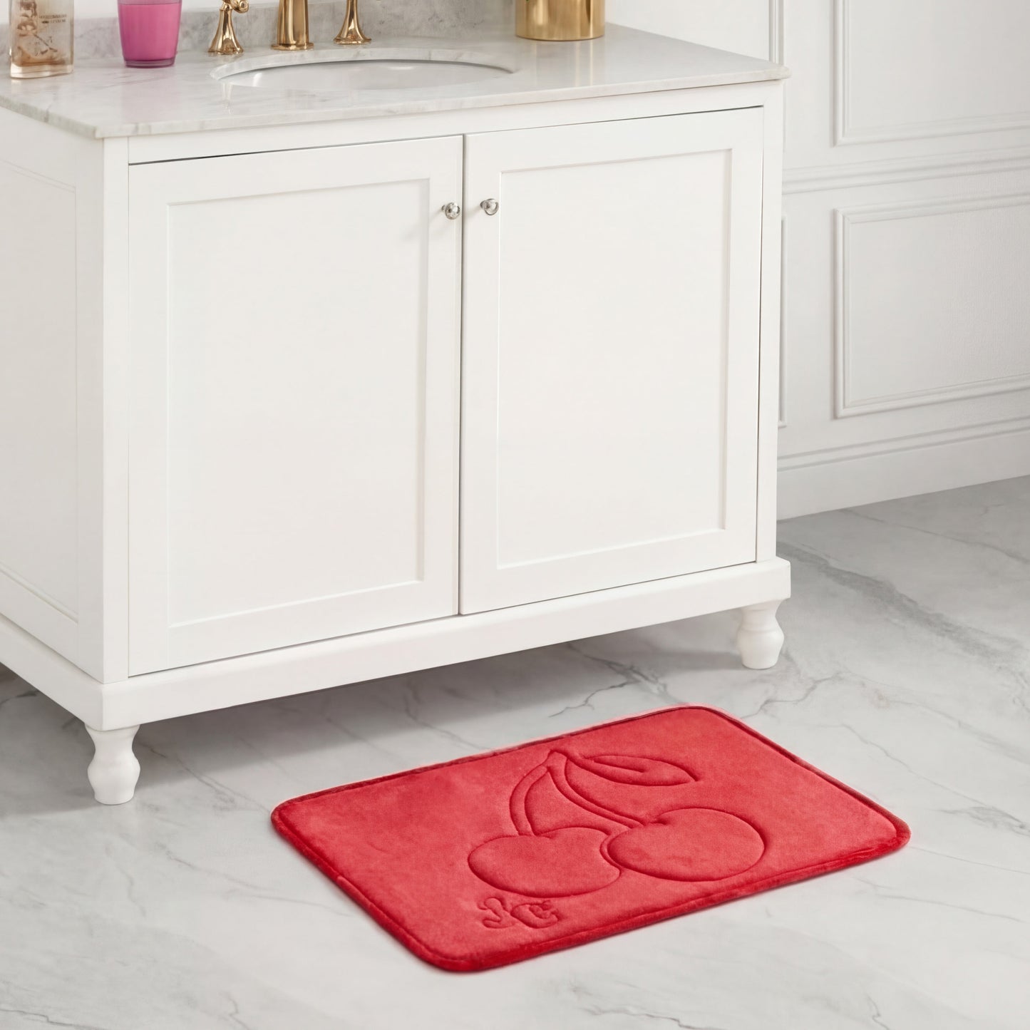A Quilted Memory Foam Bath Mat by Juicy Couture, red with an embossed cherry design, sits on white tile in front of a double-door white bathroom cabinet with a marble top.