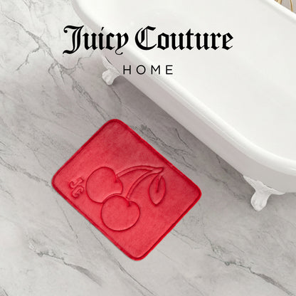 The Quilted Memory Foam Bath Mat by Juicy Couture lies on a marble floor near a white clawfoot tub. In red with embossed cherries and JC initials, this luxury mat adds comfort and style. "Juicy Couture HOME" is displayed at the top.