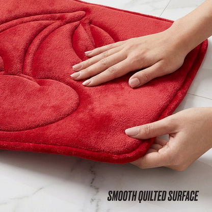 Two hands lift a red Quilted Memory Foam Bath Mat featuring a raised cherry design. The mat sits on a white marble surface. Text below reads: SMOOTH QUILTED SURFACE.