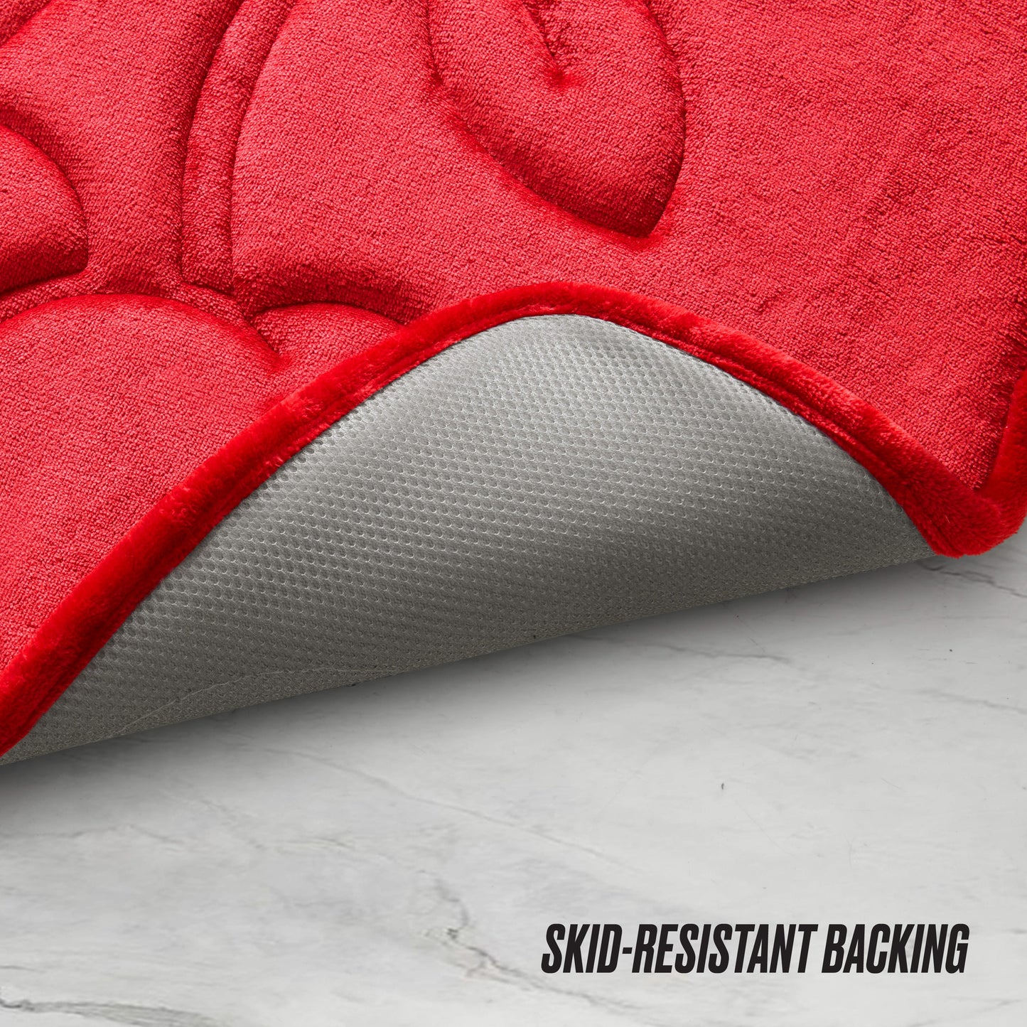 The Quilted Memory Foam Bath Mat in red is pictured partially flipped to display its gray skid-resistant backing on a white marble floor, with “SKID-RESISTANT BACKING” in black text at the bottom right.