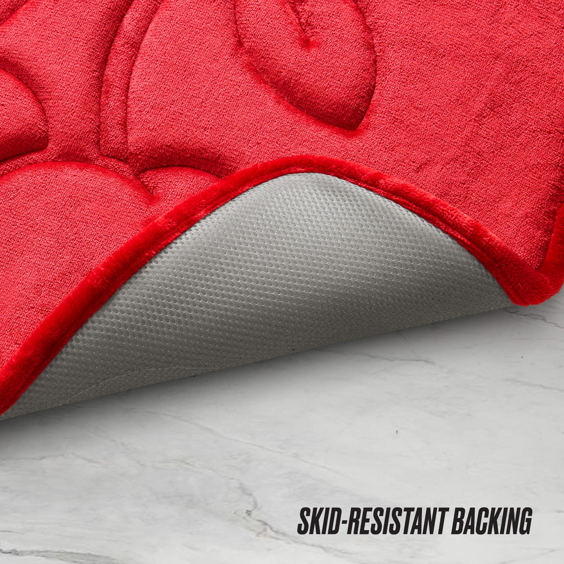 The Quilted Memory Foam Bath Mat in red is pictured partially flipped to display its gray skid-resistant backing on a white marble floor, with “SKID-RESISTANT BACKING” in black text at the bottom right.