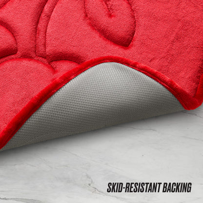 The Quilted Memory Foam Bath Mat in red is pictured partially flipped to display its gray skid-resistant backing on a white marble floor, with “SKID-RESISTANT BACKING” in black text at the bottom right.