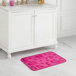 A pink Quilted Memory Foam Bath Mat with embossed bows, hearts, and flowers is placed on a white marble bathroom floor in front of a white vanity cabinet with gold faucet fixtures.