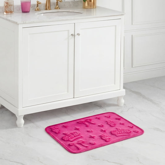 A pink Quilted Memory Foam Bath Mat with embossed bows, hearts, and flowers is placed on a white marble bathroom floor in front of a white vanity cabinet with gold faucet fixtures.