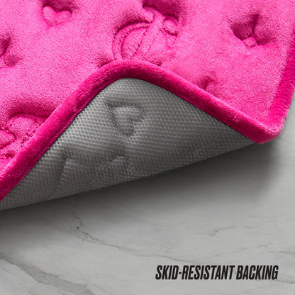 A close-up of the Quilted Memory Foam Bath Mat in pink, featuring heart-shaped quilting and a gray skid-resistant backing on a marble floor. The text SKID-RESISTANT BACKING appears in the bottom right corner.