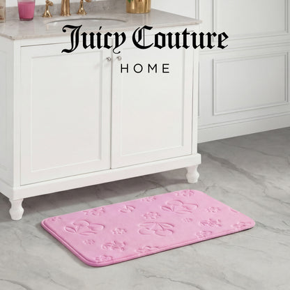 A Quilted Memory Foam Bath Mat in pink with embossed floral and bow designs lies on a marble bathroom floor by a white vanity. The skid-resistant mat adds safety, while Juicy Couture HOME is displayed at the top of the image.