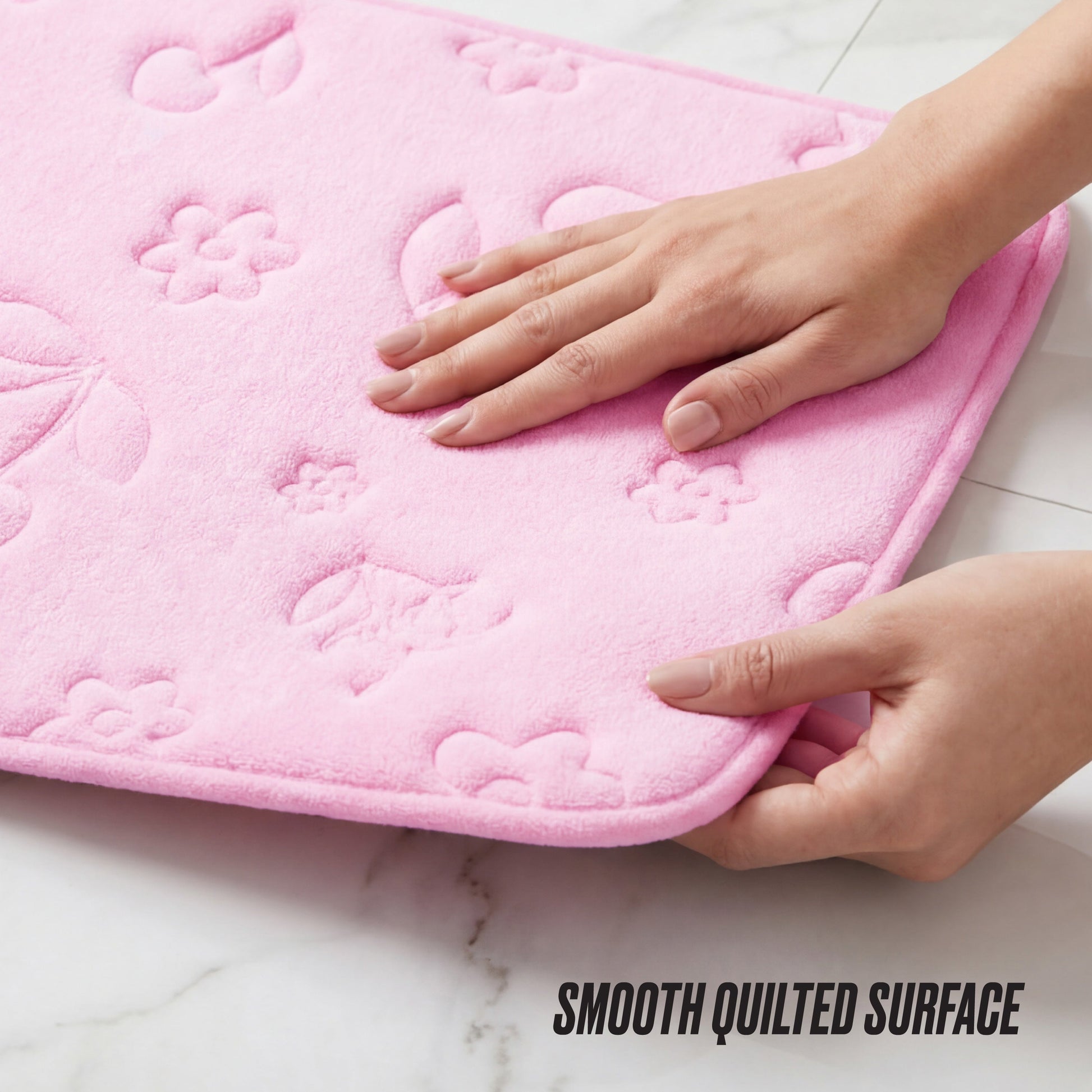 Two hands hold a pink Quilted Memory Foam Bath Mat, featuring an embossed floral and butterfly pattern, on a white tiled floor. The text "SMOOTH QUILTED SURFACE" appears in the lower right corner.