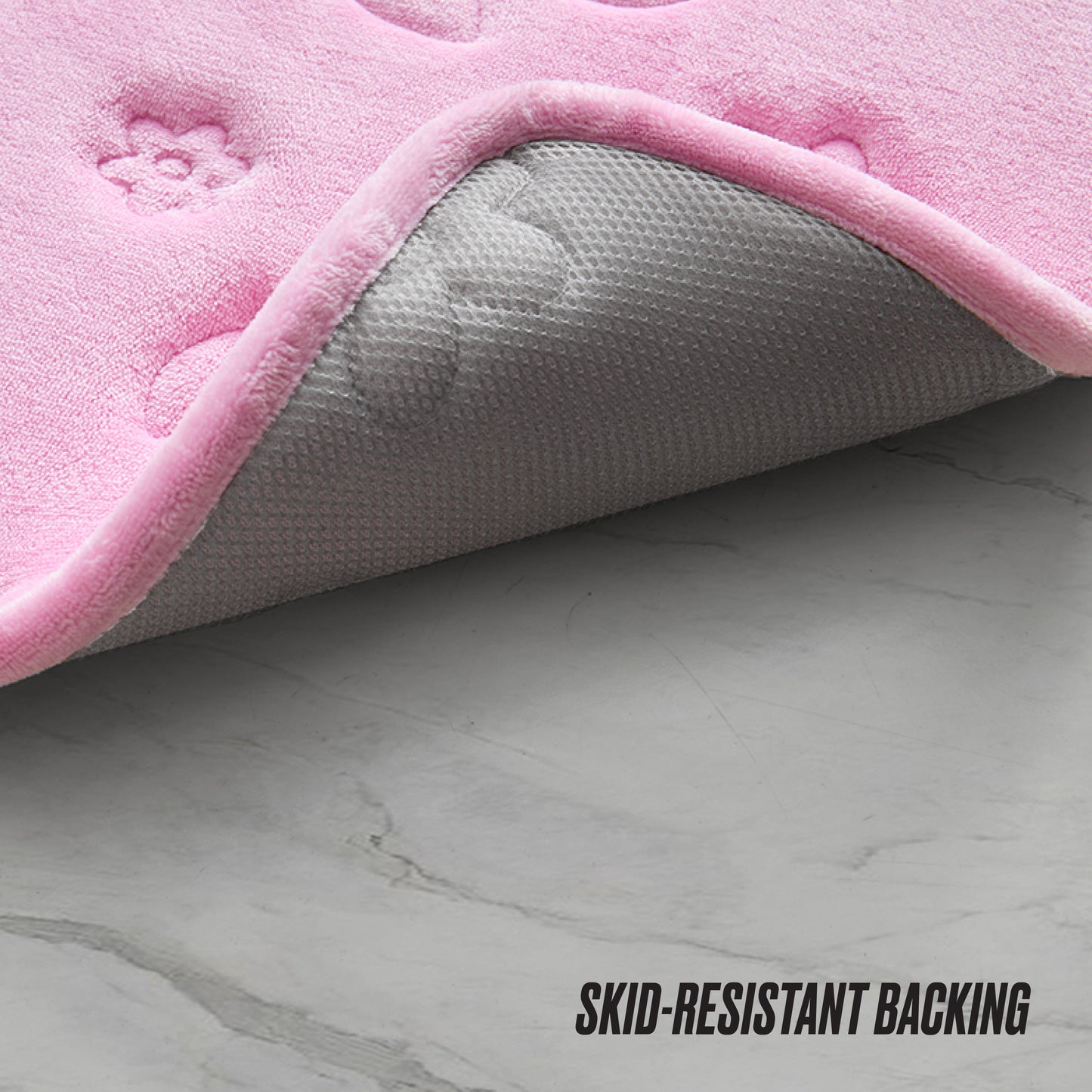A close-up of a pink Quilted Memory Foam Bath Mat with embossed paw prints, partially lifted to reveal its gray, textured skid-resistant backing on a marble floor. Text reads “SKID-RESISTANT BACKING.”.