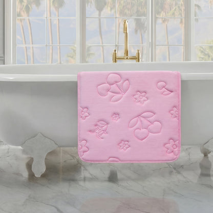 A Quilted Memory Foam Bath Mat in pink with cherry and flower designs hangs over a white clawfoot tub in a bright bathroom featuring marble floors and large windows with views of palm trees.