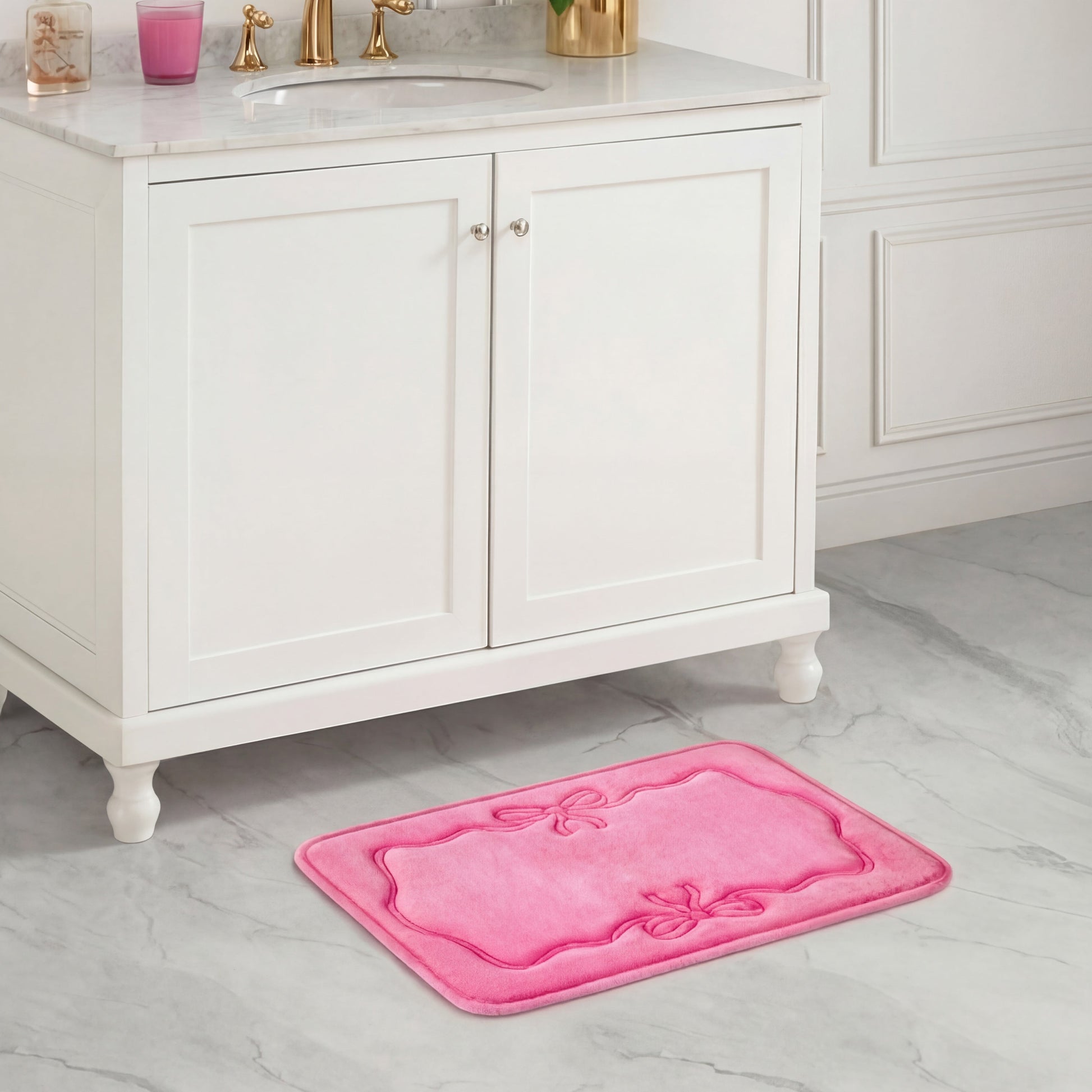 A Quilted Memory Foam Bath Mat, 17" x 24", in plush pink with decorative bows sits on a gray marble floor, complementing the white vanity and gold faucets.